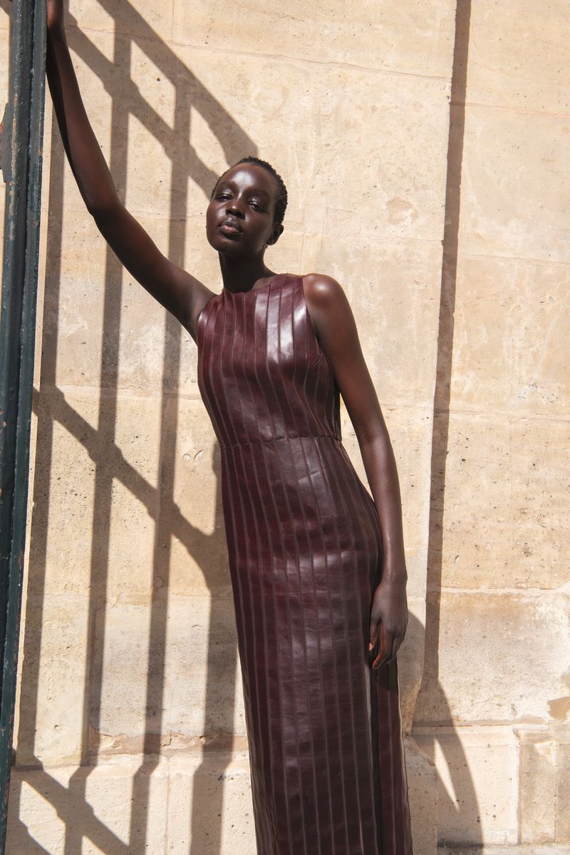 A striking image features a Black female fashion model in a sleeveless, full-length maroon leather dress, leaning against a textured wall.
