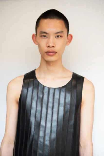 An Asian man with a shaved head is shown from the chest up wearing a black tank top with vertical seams, against a white background.