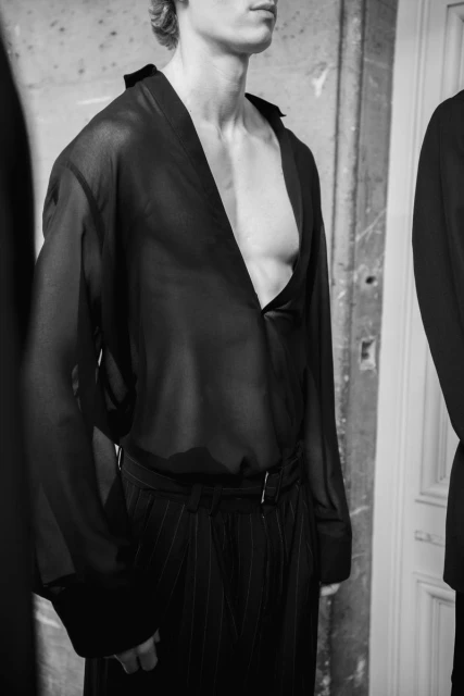 Black and white fashion shot of a slim male model in a sheer, dark top and pinstripe pants standing against a weathered wall indoors.