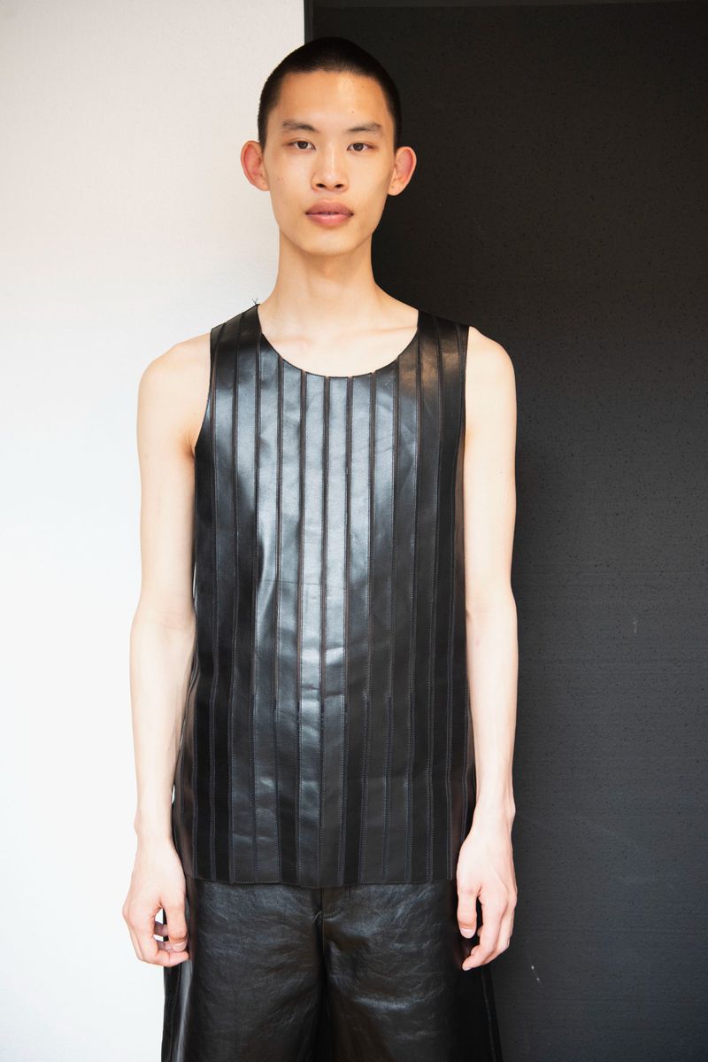 Portrait of a young adult male with short hair, wearing a black leather tank top and shorts, standing indoors against a two-tone wall.