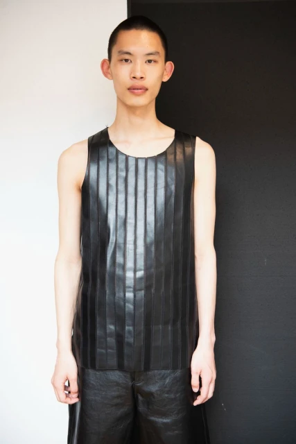 Portrait of a young adult male with short hair, wearing a black leather tank top and shorts, standing indoors against a two-tone wall.