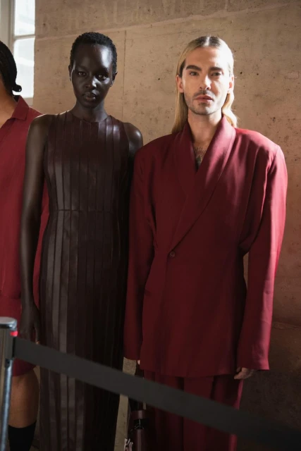 Runway fashion show featuring two models in front of a textured wall wearing burgundy and brown clothing in an indoor setting.