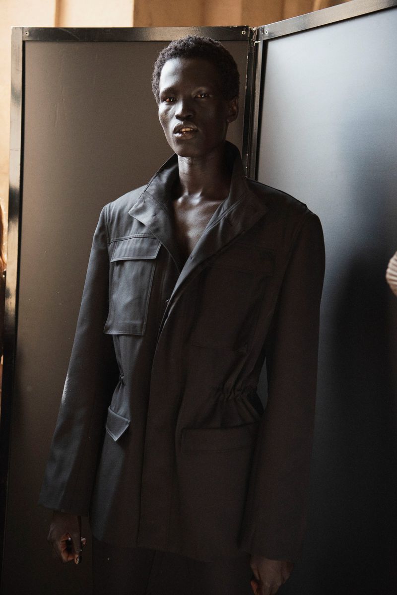 Fashion shot of a Black male model in a dark jacket standing in an indoor studio with dark walls and bright light.
