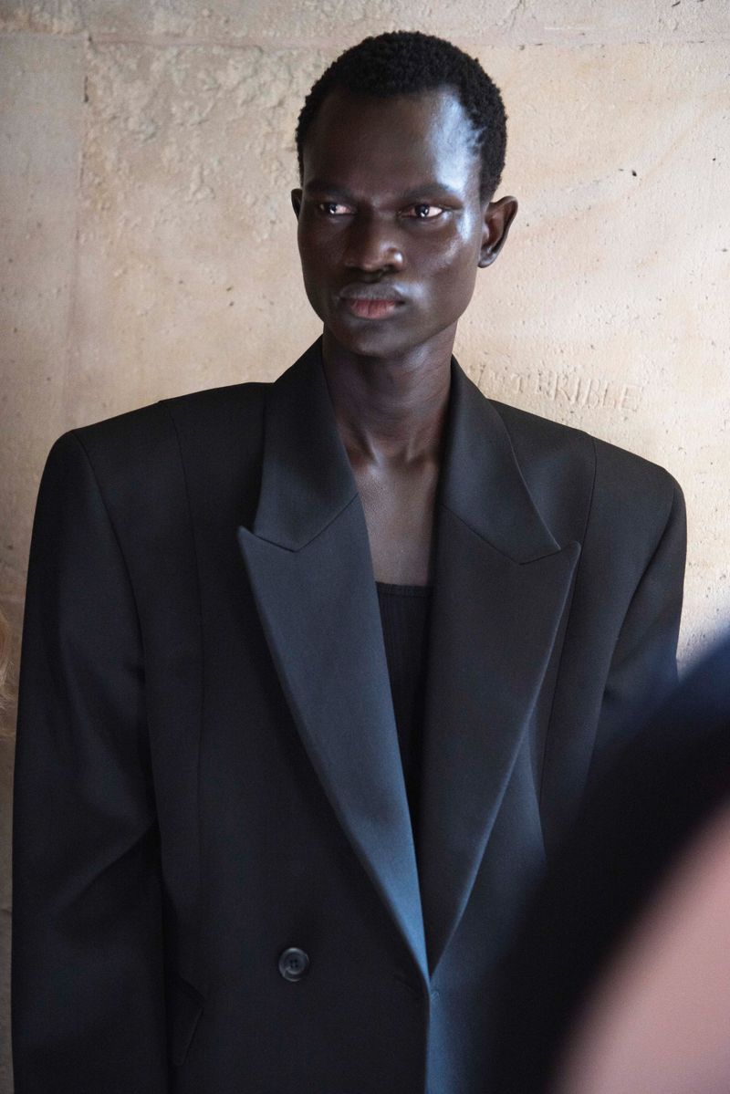 Portrait of a black male fashion model wearing a large black double-breasted blazer against a neutral textured wall backdrop.