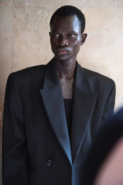 Portrait of a black male fashion model wearing a large black double-breasted blazer against a neutral textured wall backdrop.