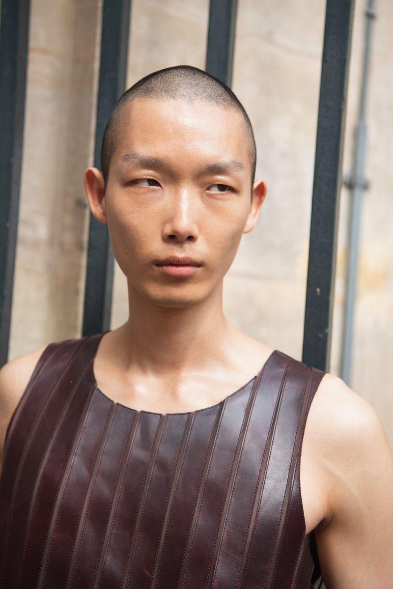 Portrait of a young Asian man with a shaved head wearing a sleeveless, maroon leather shirt, standing against a textured wall.