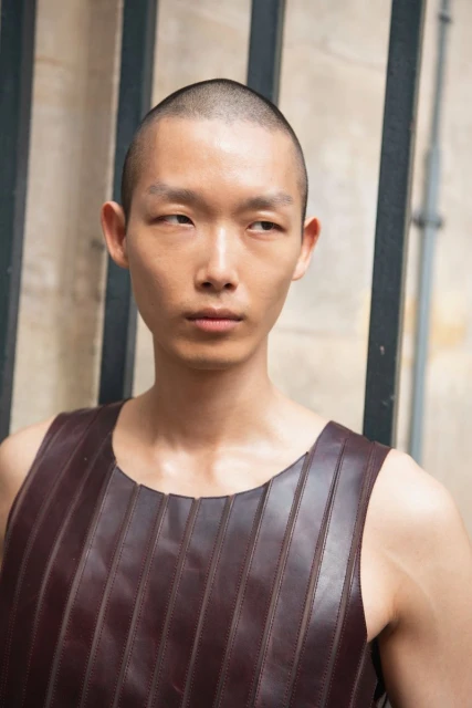 Portrait of a young Asian man with a shaved head wearing a sleeveless, maroon leather shirt, standing against a textured wall.