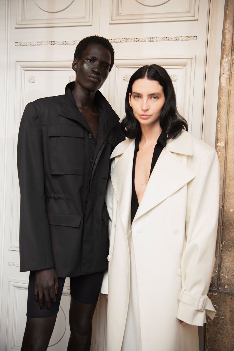 Two fashion models wearing coats pose indoors against an ornate door. One model wears black, the other wears white in a studio setting.