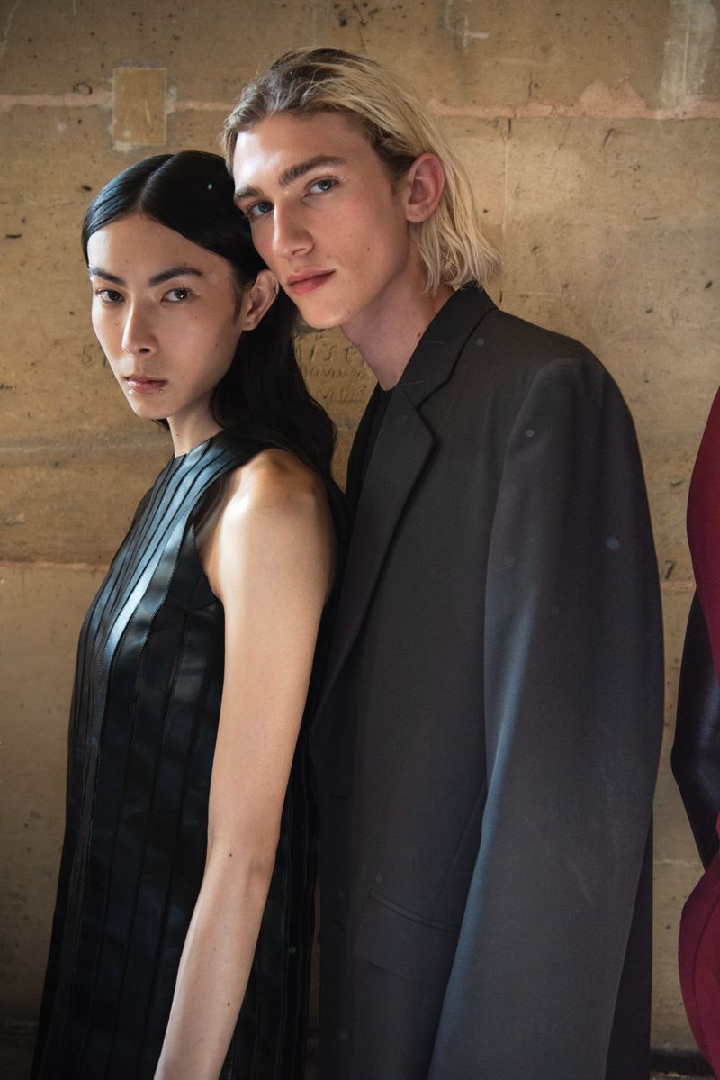 An Asian woman and a blonde male fashion model pose together in a photo studio wearing a black dress and a black suit.