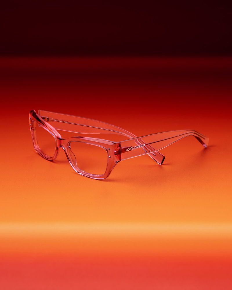 Pink-tinted glasses on a gradient background transitioning from warm orange to deep burgundy. Product shot highlights frame details.