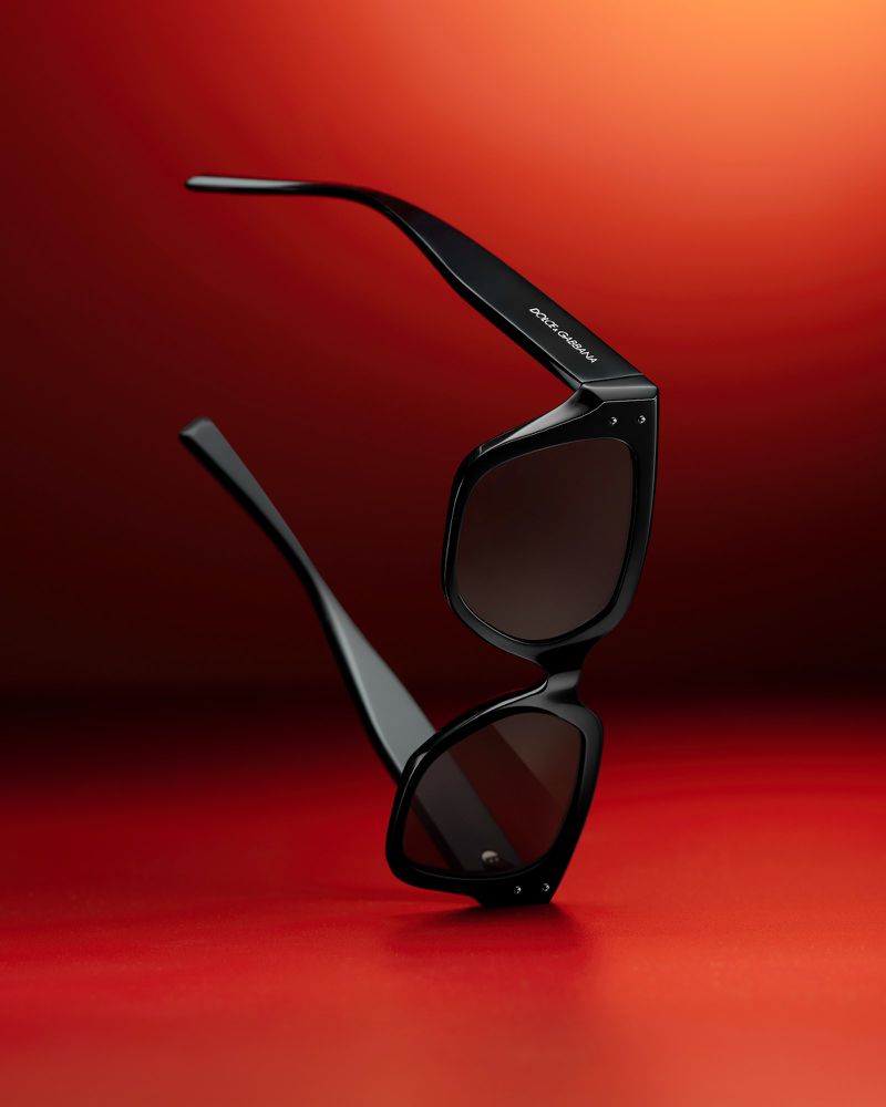 Black Dolce & Gabbana sunglasses on a red reflective surface, featuring a sleek design and highlighting the brand's logo on the temple.