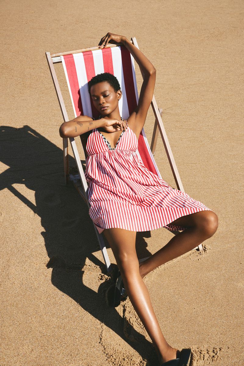 A woman in a red-and-white striped dress rests on a striped beach chair on the sand under sunlight, casting a shadow. Summer fashion.