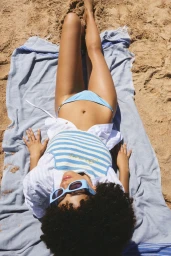 Model lying on a blue striped towel on a sandy beach wearing a blue and white striped top and sunglasses.