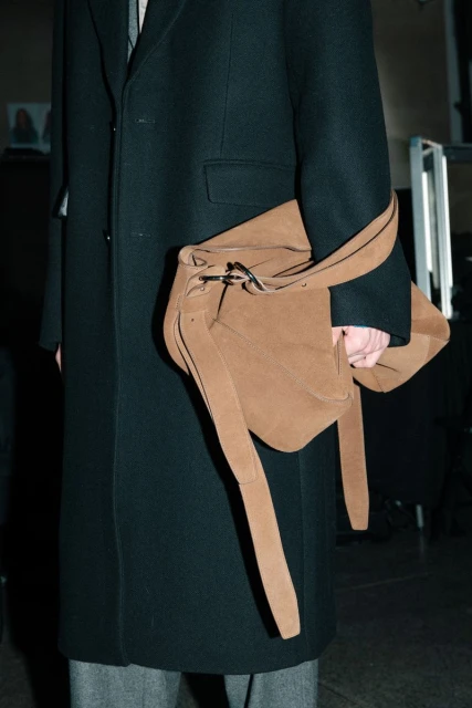 Person in black overcoat holding a tan suede bag with long tassels. The scene is indoors with soft lighting and neutral tones.