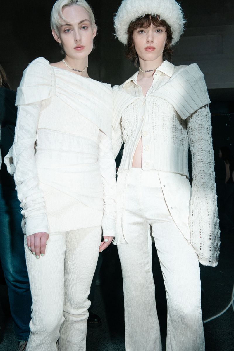 Two women in matching white outfits and a white hat posing in a studio or indoor fashion setting; both wear subtle necklaces.