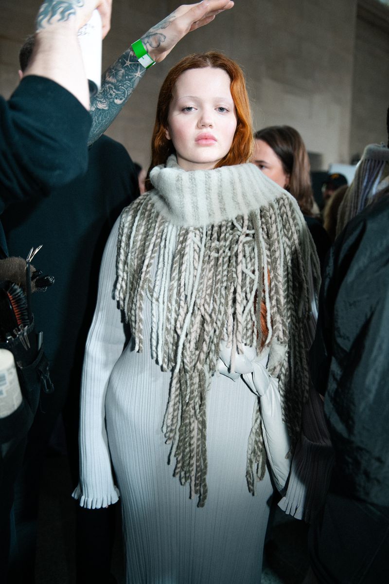 Fashion shot: a pale-skinned woman with red hair wears a light gray dress and fringed shawl, posing indoors amongst a crowd, with muted lighting.