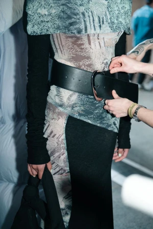 Runway fashion model wearing a patterned top and black skirt is styled with a wide black belt and adjusted by two fashion stylists.