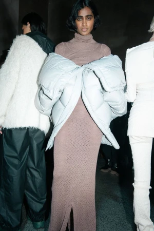 Fashion shoot featuring a female model in a brown turtleneck dress with an oversized jacket, backstage with other models.