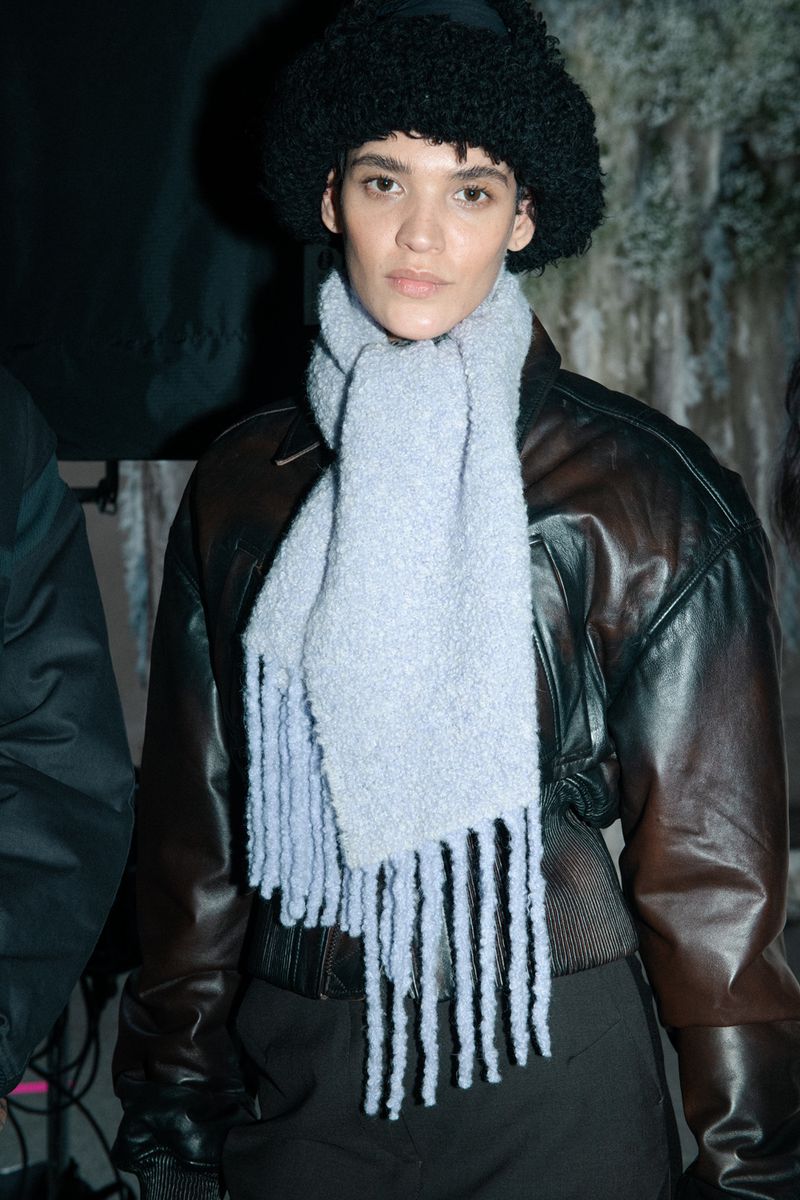 Woman in a black hat and light blue scarf poses in front of mottled backdrop, wearing a dark jacket. Fashion and style.