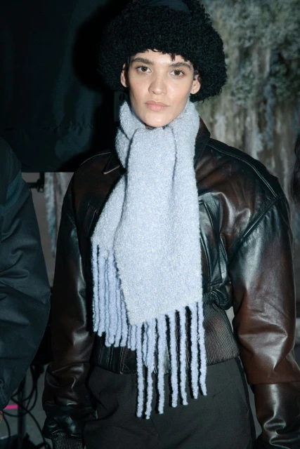Woman in a black hat and light blue scarf poses in front of mottled backdrop, wearing a dark jacket. Fashion and style.