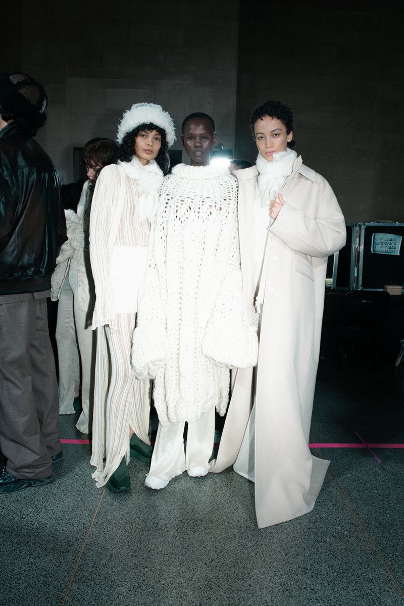 Three female fashion models posing indoors, wearing white dresses and coats, against a neutral background. Fashion shoot, studio lighting.