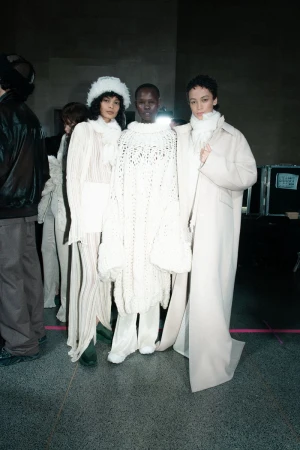 Three female fashion models posing indoors, wearing white dresses and coats, against a neutral background. Fashion shoot, studio lighting.
