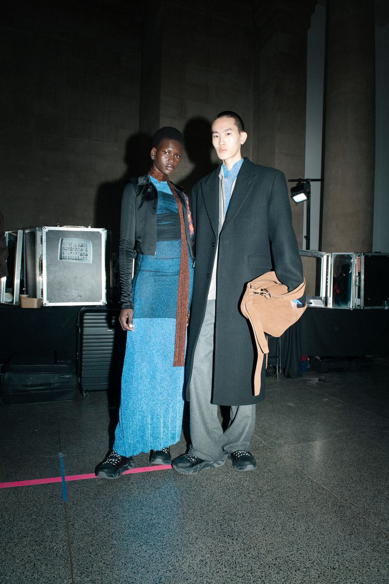 Two fashion models pose in a dimly lit backstage setting, the woman wears a shimmering blue dress, and the man a black coat with beige bag.