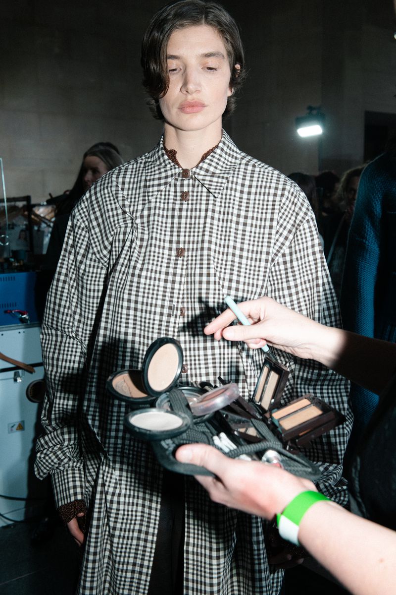 Woman in a checkered shirt having her makeup applied indoors by an artist with a makeup palette, preparing for a fashion show.