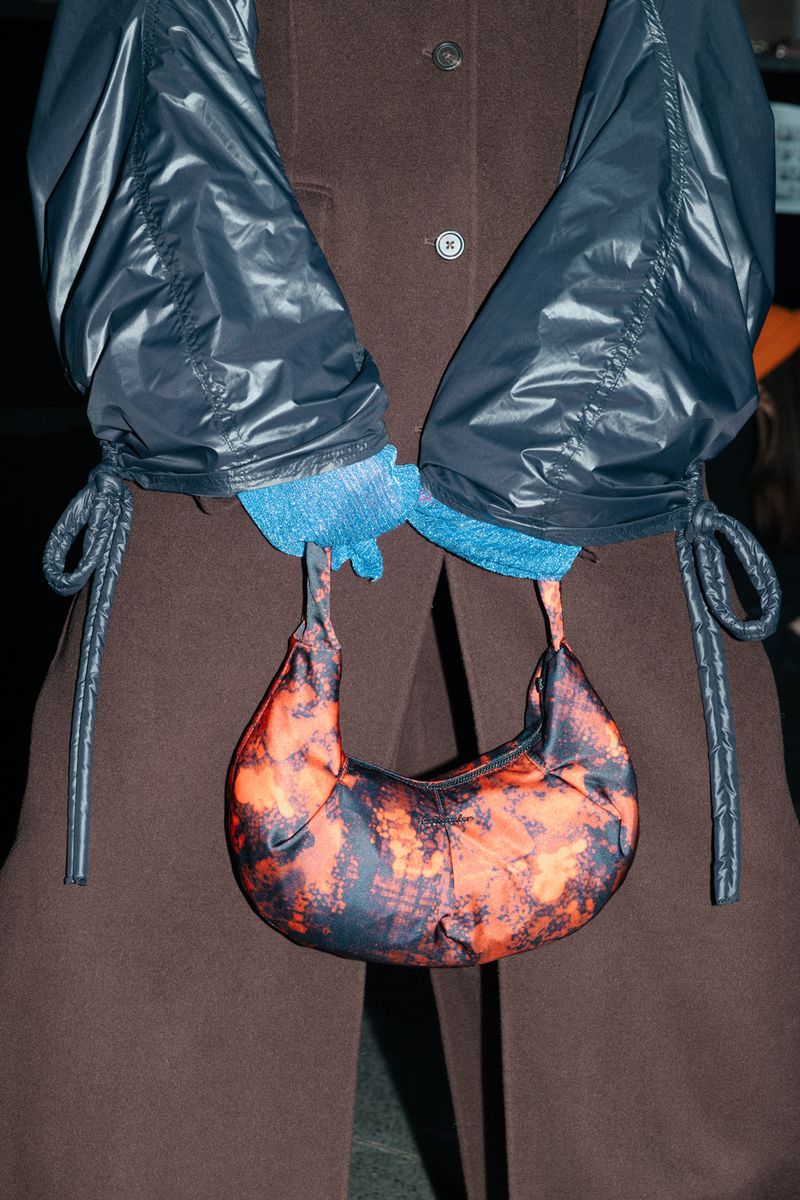 A person in a brown coat holds an orange and black crescent-shaped bag with a blue-gloved hand and black nylon jacket sleeves.