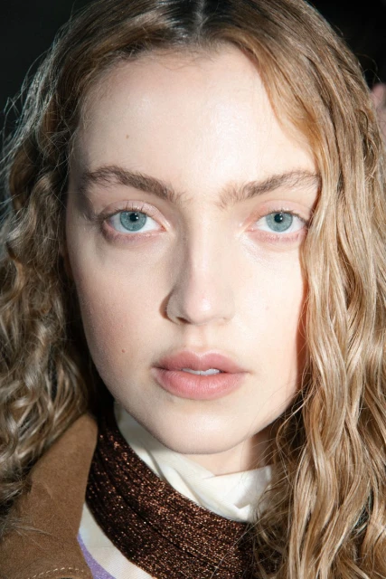 Close-up portrait of a young adult woman with light skin, light blue eyes, and wavy blonde hair, wearing a tan jacket indoors.
