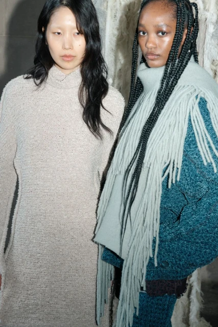 Two women pose in a studio, one with long braids and a scarf with fringe, the other with long loose dark hair and a knit dress.