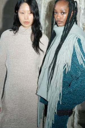 Two women pose in a studio, one with long braids and a scarf with fringe, the other with long loose dark hair and a knit dress.