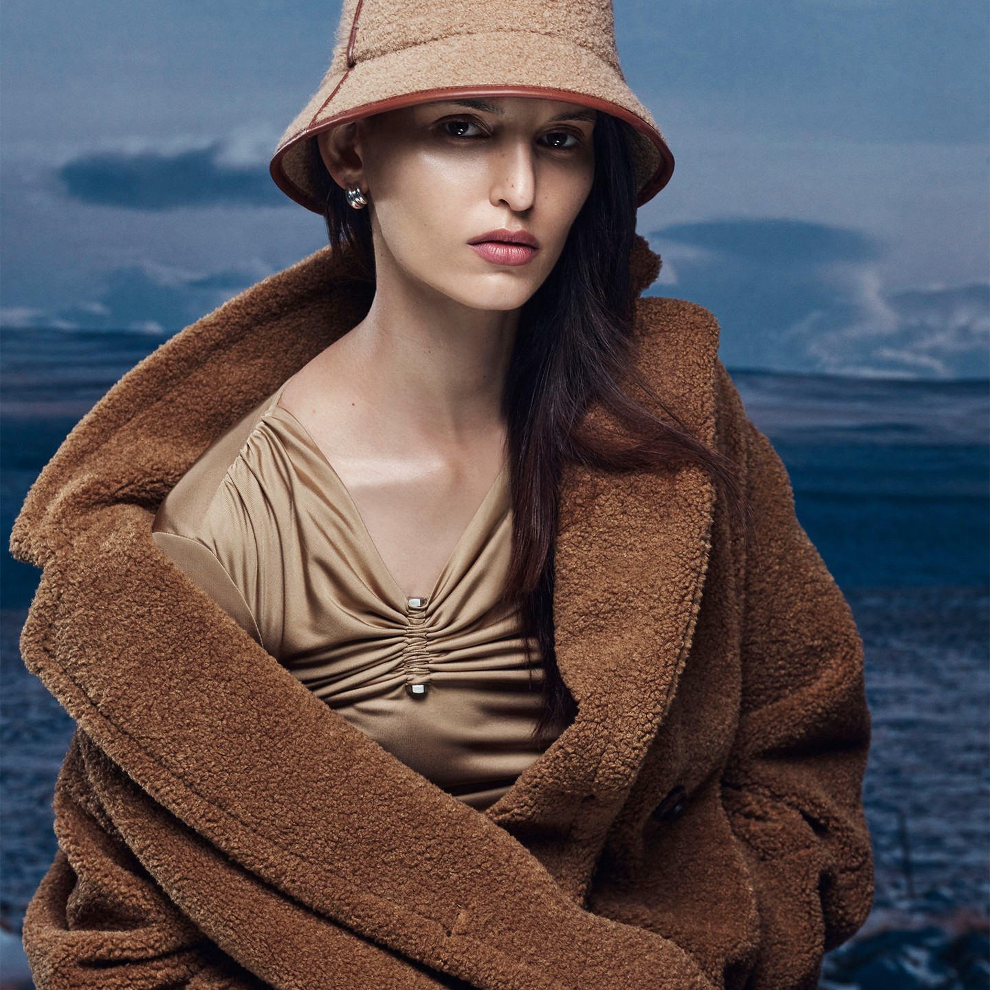 Portrait of a woman in a beige bucket hat and brown coat against a diffused blue background with a horizon line.