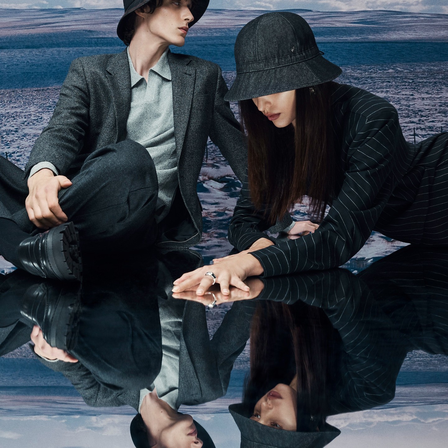 A stylish couple poses in matching bucket hats and suits, reflecting off a glossy surface against a wintry, blue-toned landscape backdrop.