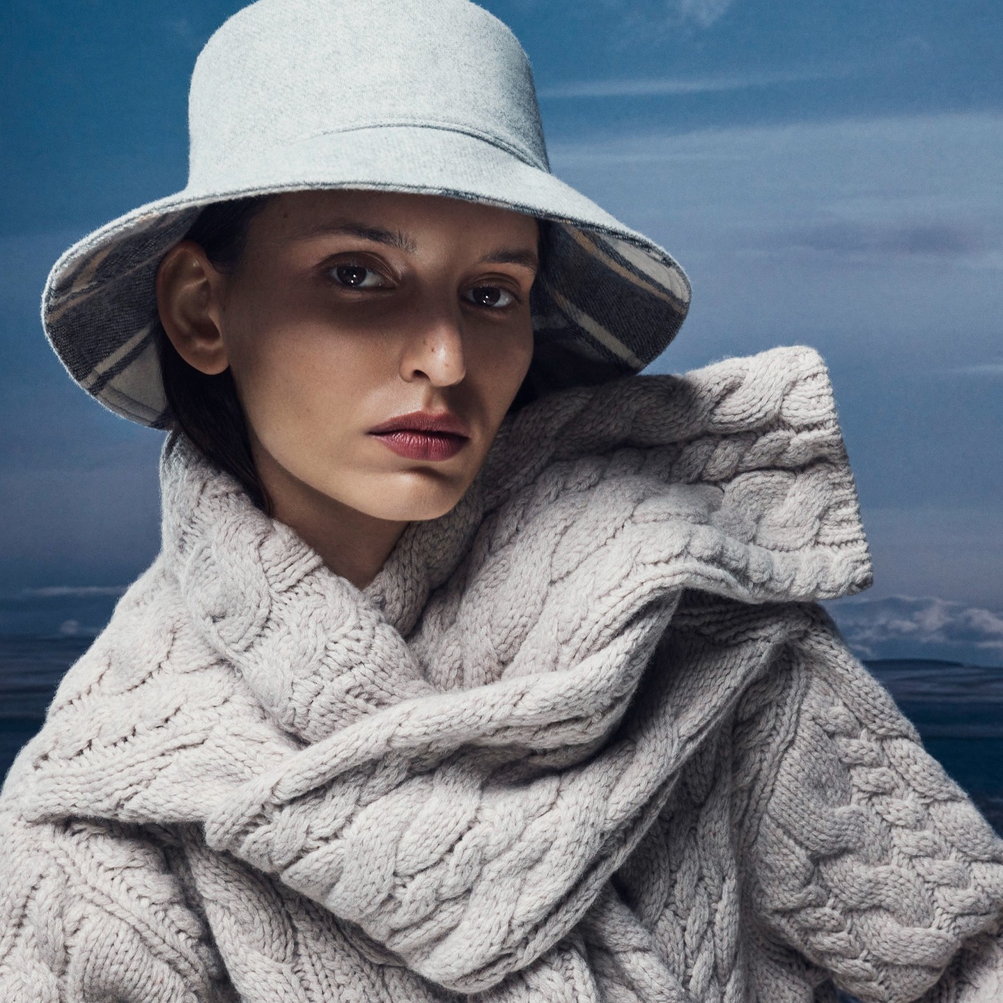 Close-up of woman wearing a knit scarf and bucket hat against a cloudy sky. The woman is looking directly at the camera.