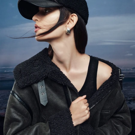 Portrait of a dark-haired woman wearing a black cap and aviator jacket against a dramatic sky. Fashion shoot, outdoor setting.