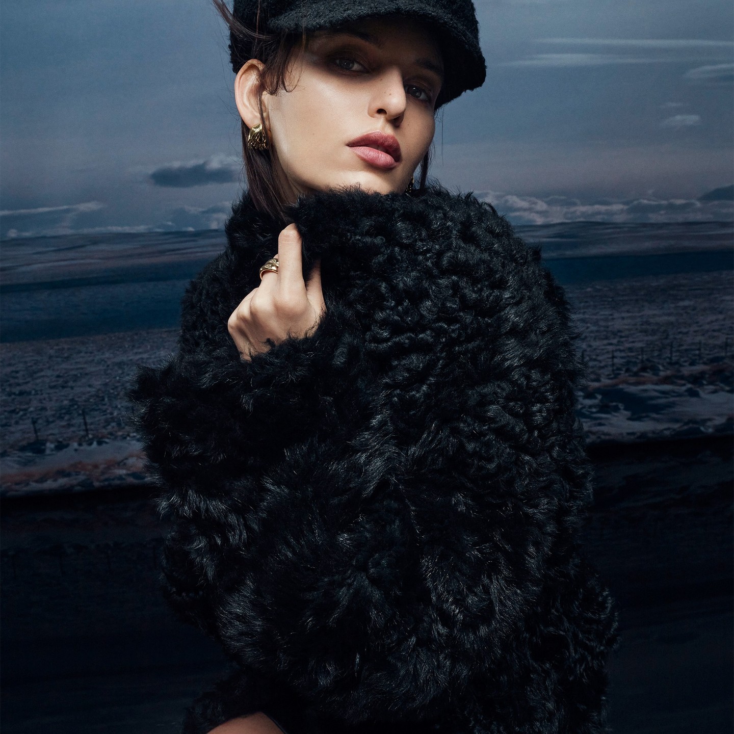 Model wearing a black fluffy textured coat and matching cap against a moody landscape background for Helen Kaminski