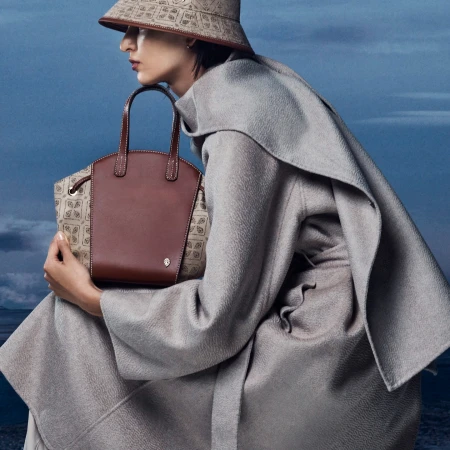 Fashionable adult female poses outdoors in a light gray coat and patterned bucket hat, holding a brown leather and tan logo handbag.