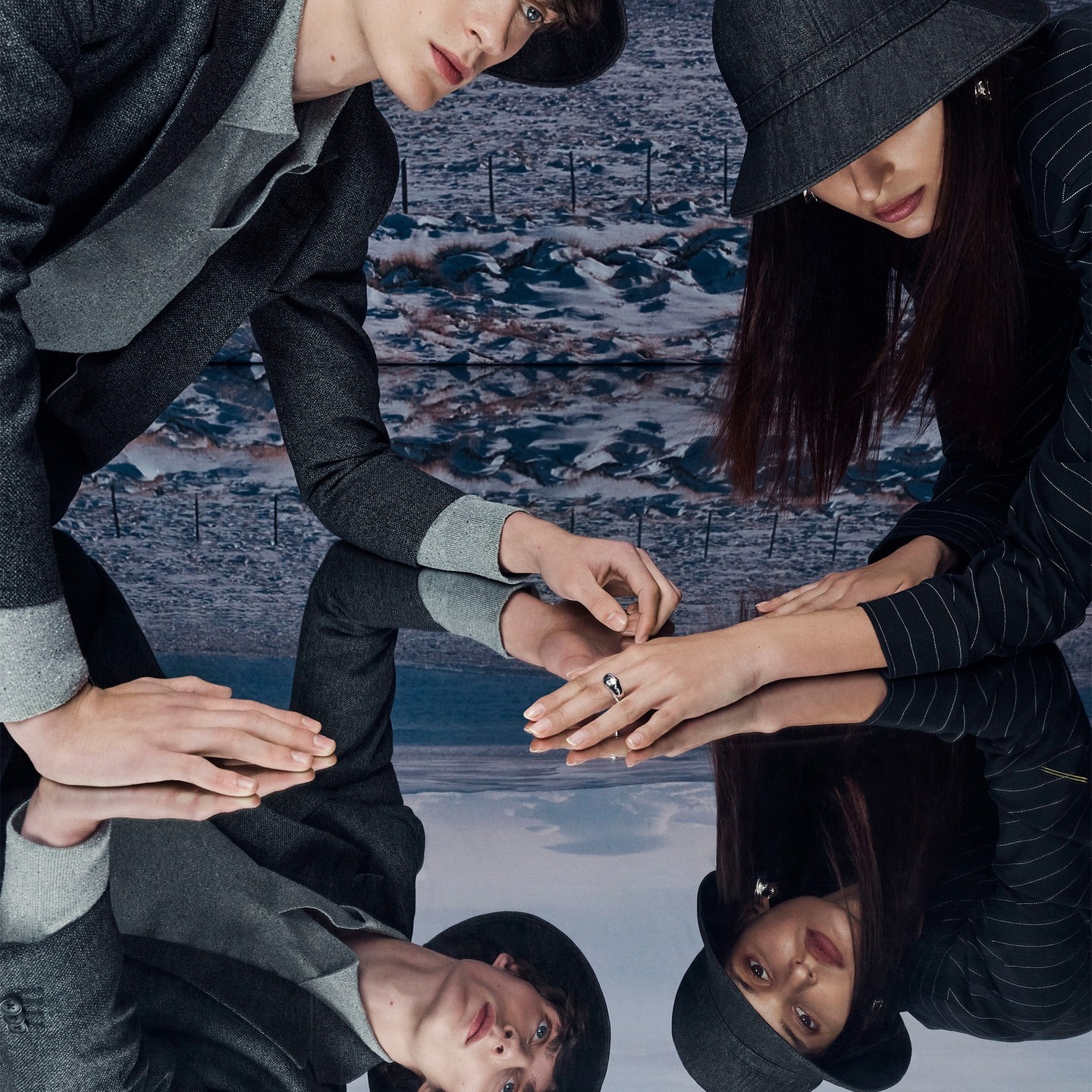 Two fashion models wearing matching hats and grey and black outfits are positioned in front of a mirrored surface and abstract background.