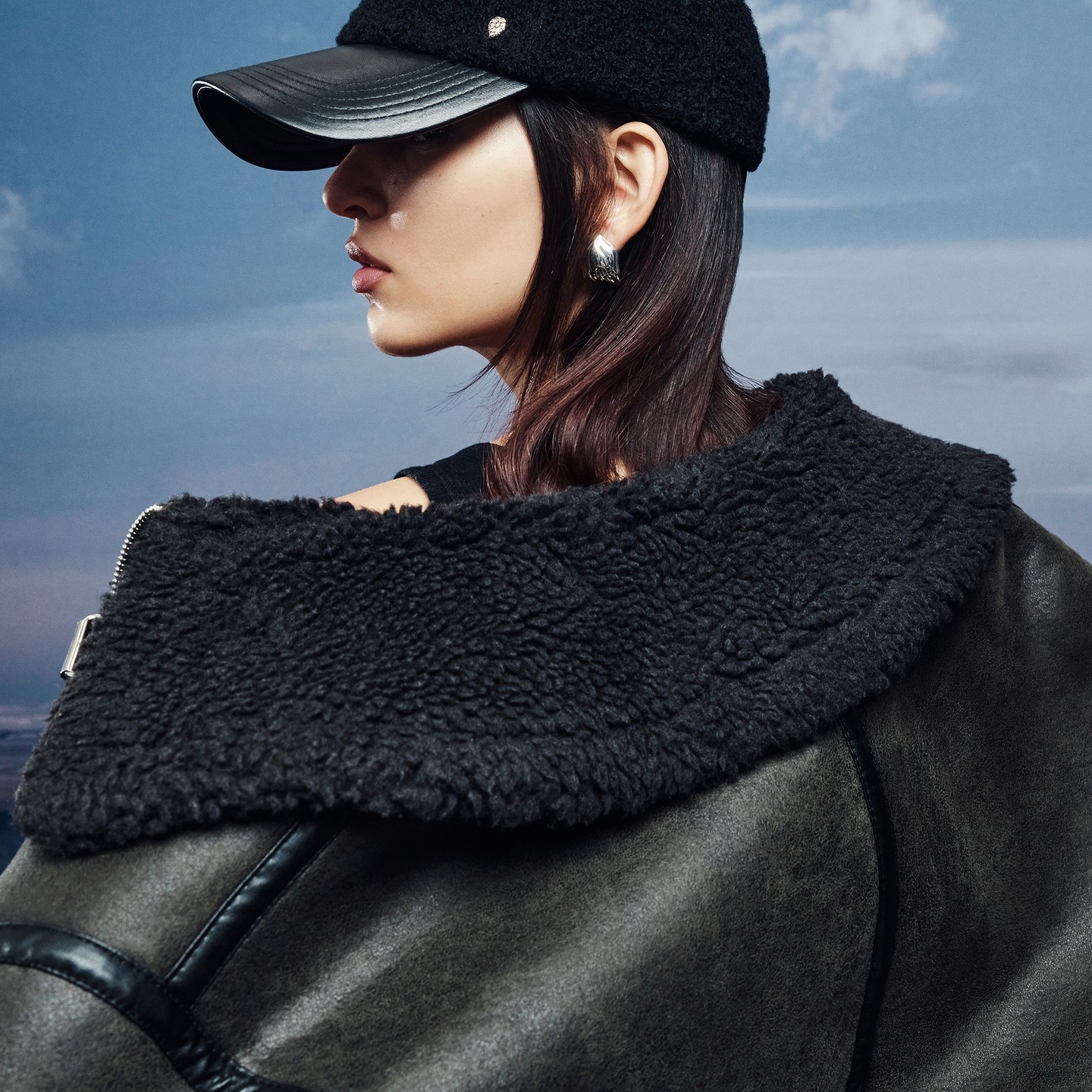 Woman in a black cap and a dark coat with a shearling collar looking over her shoulder against a blue cloudy sky background.