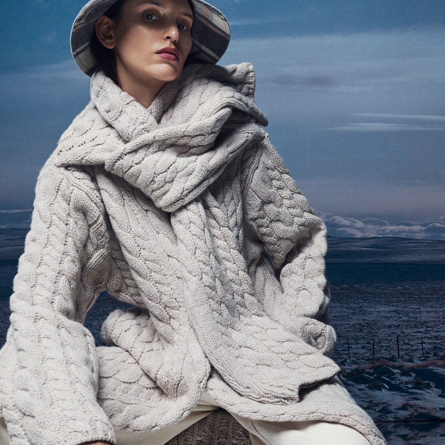 Woman wearing a grey cable-knit cardigan, scarf, and bucket hat against an outdoor backdrop of a cloudy sky above a snow-covered landscape.