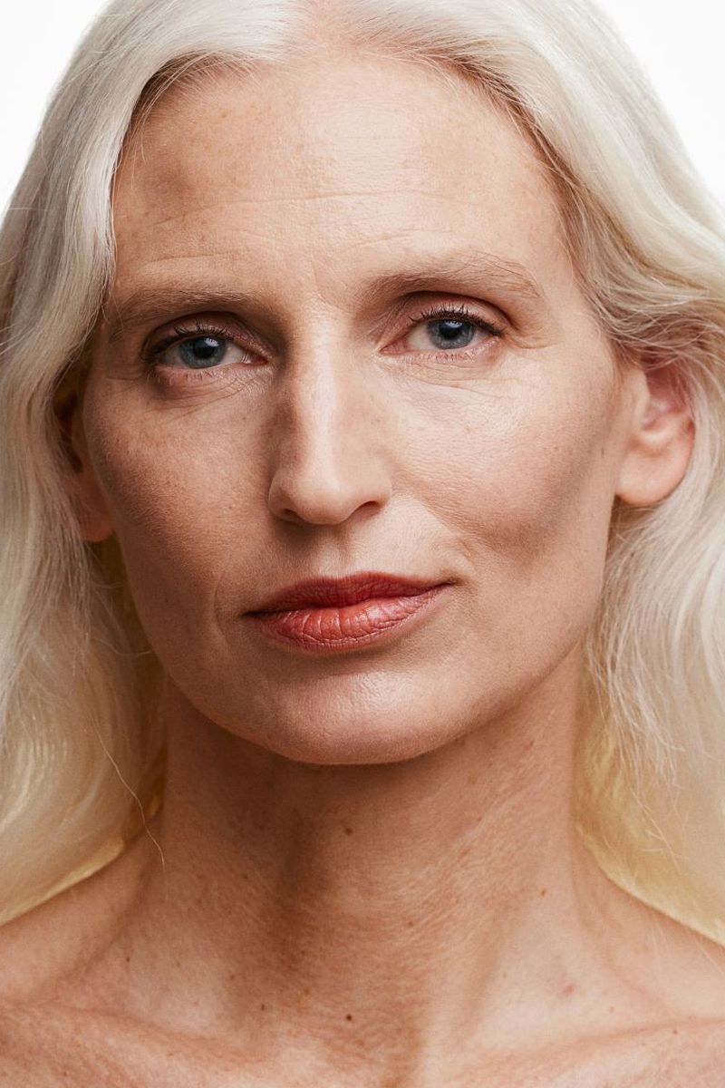 Close-up portrait of an adult woman with light skin, blue eyes, and long white hair, looking directly at the camera against a white background.