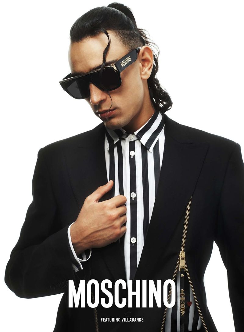 A man in sunglasses and a striped shirt wearing a black suit jacket promotes the Moschino brand against a solid white backdrop.