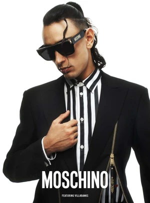 A man wearing Moschino sunglasses and a black blazer with zipper details against a white background.