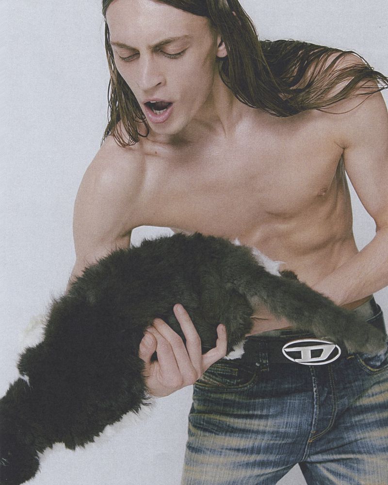 A shirtless male model holds a fluffy black and white cat. The man is wearing jeans with a Diesel brand belt and has long brown hair.