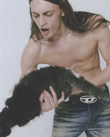 A shirtless male model holds a fluffy black and white cat. The man is wearing jeans with a Diesel brand belt and has long brown hair.