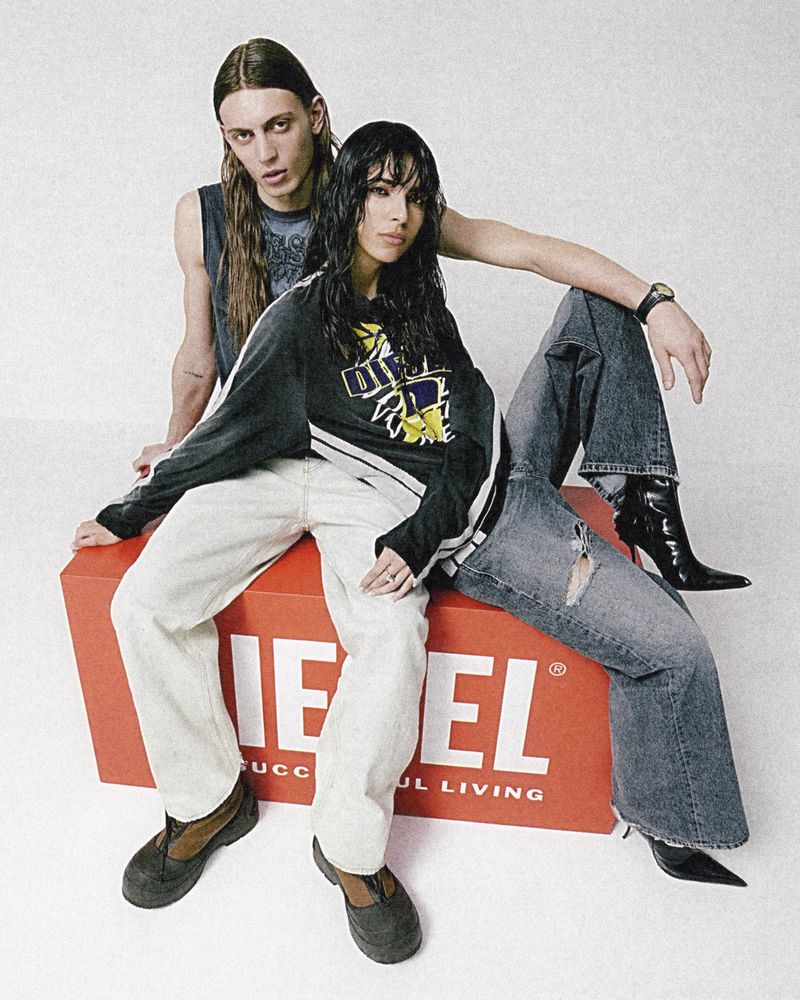 Two fashion models pose in Diesel apparel, sitting on a red cube logo. The male model wears a tank top, and the female wears a sweatshirt and jeans.