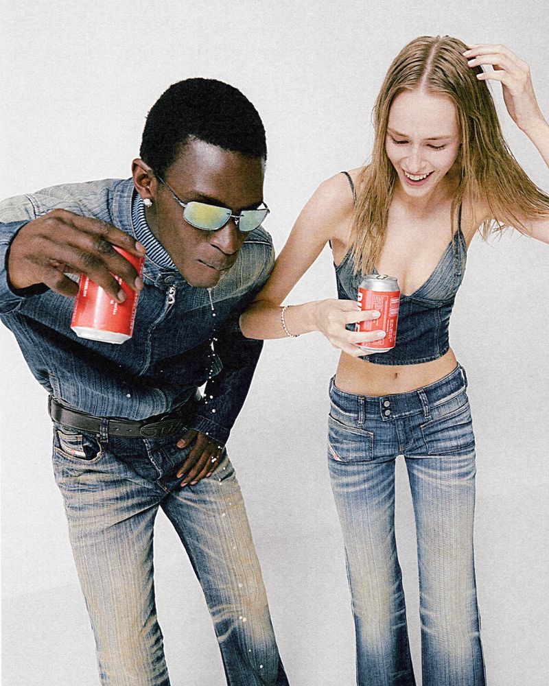 Two models, one Black and one White, pose in denim outfits holding red cans. The male model spills his drink against a white backdrop.