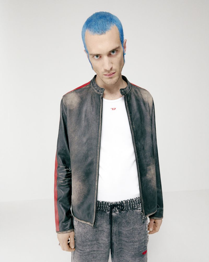 Adult male poses indoors wearing a black moto jacket, white tee-shirt, and grey sweatpants against a bright white backdrop.