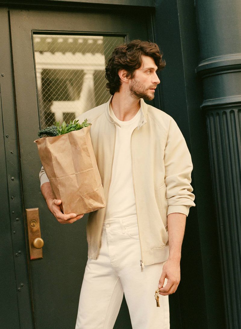 A dark-haired man in a jacket carries a brown paper grocery bag outside against a dark green door on a well-lit day. 
